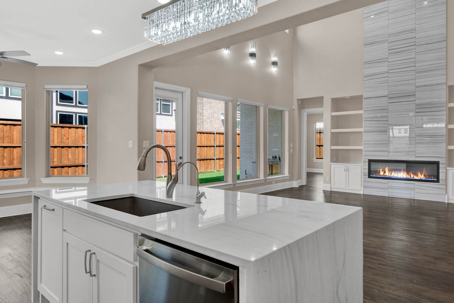 1233 Zulaya_FB-13 Modern kitchen with a large white island, double sink, and dishwasher. Spacious living area features floor-to-ceiling windows, a sleek fireplace, and built-in shelves.